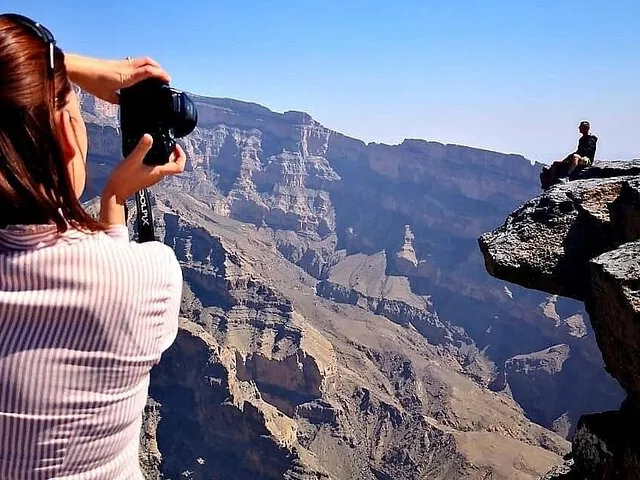 Donna che scatta una foto a una persona seduta sul ciglio della scogliera, affacciata su un profondo canyon.