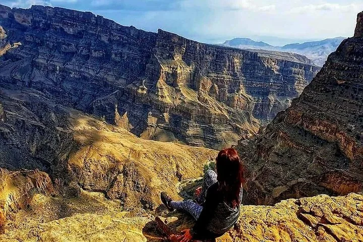 Person sitting on cliff edge looking at deep gorge and layered mountains.