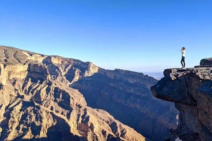 Person standing on jutting rock overlooking vast Jebel Shams canyon.