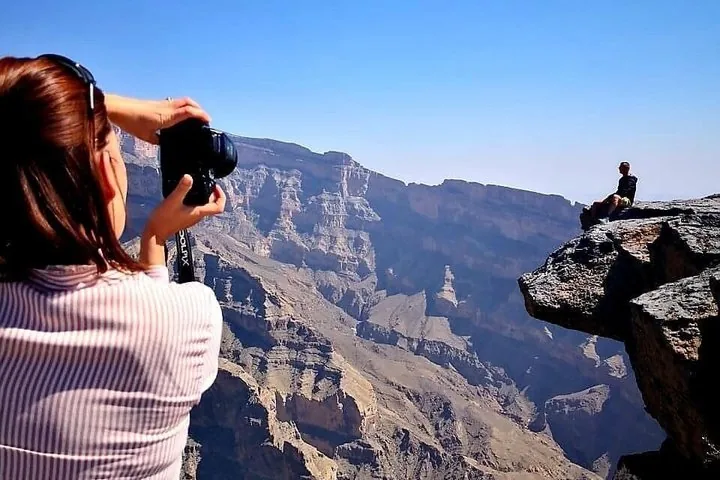 Femme prenant en photo une personne assise sur un rocher saillant au-dessus d'un canyon profond.